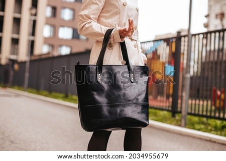 A girl in a beige raincoat is holding a large black bag made of genuine leather. Leather products. Autumn walk around the city. Black leather shopping bag Royalty-Free Stock Photo #2034955679