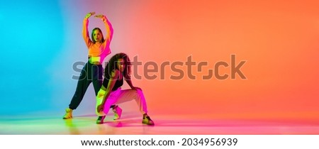 Portrait of two young beautiful hip-hop female dancers in modern clothes on colorful gradient blue orange at dance hall in neon. Youth culture, movement, active lifestyle, action, street dance, ad Royalty-Free Stock Photo #2034956939
