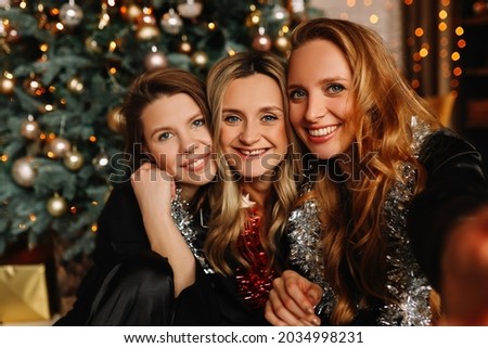 Happy funny women girlfriends in Santa hats celebrate Christmas holiday laugh take photo selfies using a mobile phone at a house party, selective focus