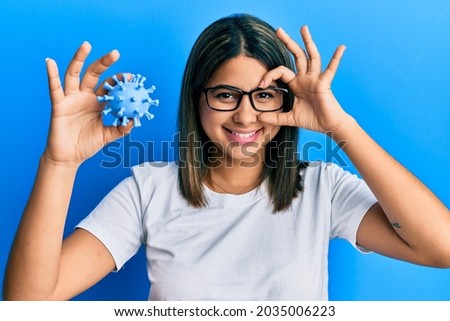 Young latin woman holding virus toy smiling happy doing ok sign with hand on eye looking through fingers 