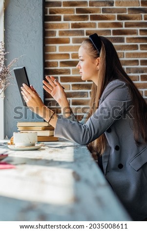 A beautiful young woman uses a tablet for video communication at a table in a cafe. The girl talks to friends. The concept of young business people working from home.