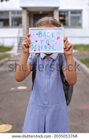 Elementary school girl in medical mask holds picture with back to school message. First offline day with social distance rules, new normal education, reunion after lockdown and quarantine