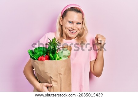 Beautiful caucasian blonde woman holding paper bag with groceries screaming proud, celebrating victory and success very excited with raised arm 