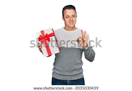 Handsome young man holding gift doing ok sign with fingers, smiling friendly gesturing excellent symbol 