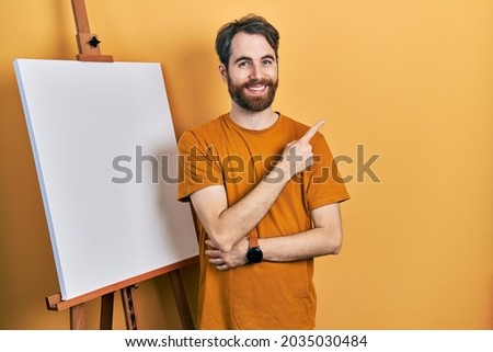 Caucasian man with beard standing by painter easel stand smiling cheerful pointing with hand and finger up to the side 