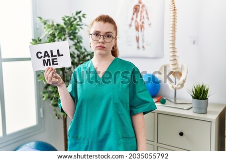 Young redhead doctor woman working at pain recovery clinic holding call me banner thinking attitude and sober expression looking self confident 