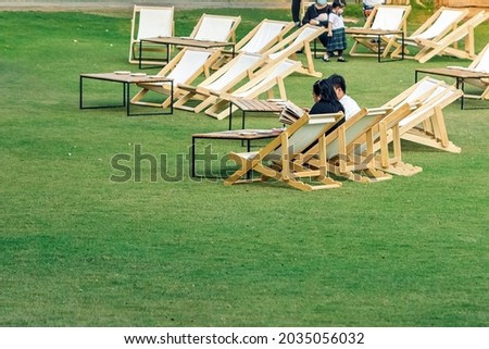 Asian young couple sit to relax and look at the menu to order food for dinner on white deck chairs with tables in lawn is surrounded by shady green grass.Comfortable on outdoor patio chairs in garden.