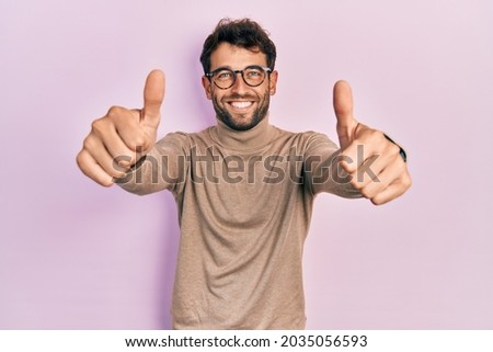 Handsome man with beard wearing turtleneck sweater and glasses approving doing positive gesture with hand, thumbs up smiling and happy for success. winner gesture. 