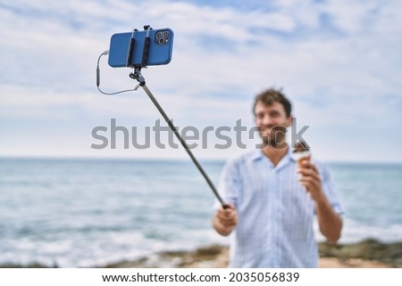 Young hispanic man eating ice cream making selfie by the smartphone at the beach.