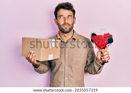 Handsome man with beard holding packing tape holding cardboard smiling looking to the side and staring away thinking. 
