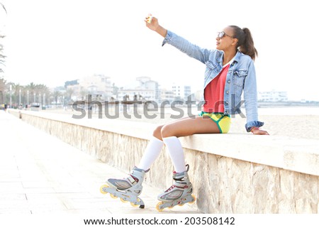 Profile view of an african american teenager with roller skates sitting by the beach in a destination city, using a smartphone to take a selfie portrait on holiday. Recreational technology lifestyle.