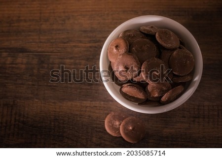 A close up shot of a small, white bowl of flat chocolate pieces or chocolate chips on a wooden kitchen table or board shot from above, overhead view