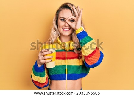 Beautiful young blonde woman drinking cup of coffee wearing headphones doing ok gesture with hand smiling, eye looking through fingers with happy face. 
