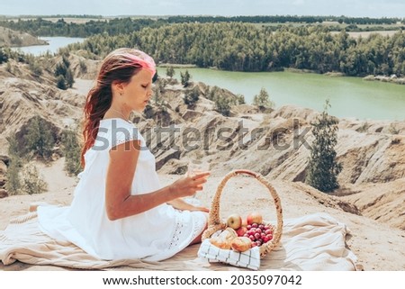 teenage girl in white dress is sitting on edge of cliff next to picnic basket and having fun during lunch, background of stunningly beautiful landscape with rocks and lake outdoor