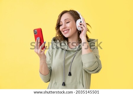 Happy teeth smile woman listen music headphones holding smartphone in hand looking screen Caucasian female enjoy podcast or audio books dressed oversize hoodie yellow background close up portrait
