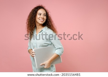 Young black african american smiling confident fun freelancer curly woman in blue shirt hold going with closed laptop pc computer look aside isolated on pastel pink color background studio portrait