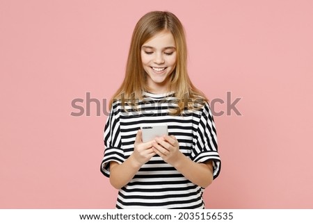 Little blonde smiling kid happy nice girl 12-13 years old wearing striped oversized t-shirt hold mobile cell phone chatting isolated on pastel pink background children. Childhood lifestyle concept