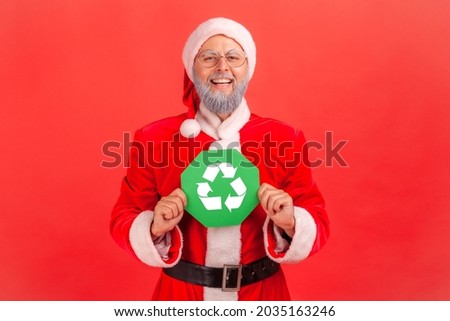 Portrait of smiling elderly man with gray beard wearing santa claus costume standing looking at camera, holding green recycling sign in hands. Indoor studio shot isolated on red background.