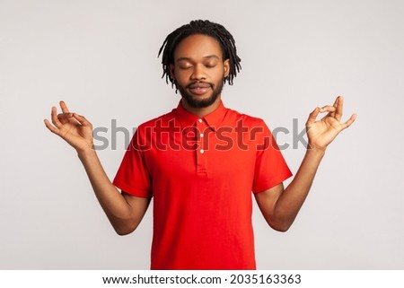 Portrait of relaxed bearded man with dreadlocks wearing red casual style T-shirt, standing with raised arms and doing yoga meditating exercise. Indoor studio shot isolated on gray background. Royalty-Free Stock Photo #2035163363