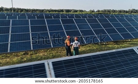 Aerial shot of three solar energy engineers on a large solar farm. Three employees of alternative power plant walking and talking about scheme of solar panels.Technician and investor