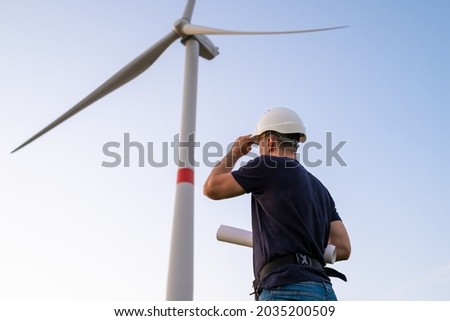 Engineer in hard hat holds a plan project for the construction of wind turbines in the field against the background of windmills. Generation of electricity. Alternative production of renewable green