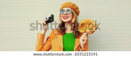 Autumn portrait of happy smiling young woman with film camera and yellow maple leaves wearing a french beret on gray background