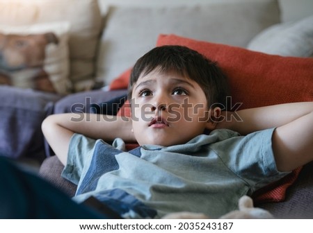 Happy boy lying on bean bag looking up watching cartoon on TV. mixed race Child resting in living room with bright light shining from window on sunny day summer. Kid relaxing at home on weekend 