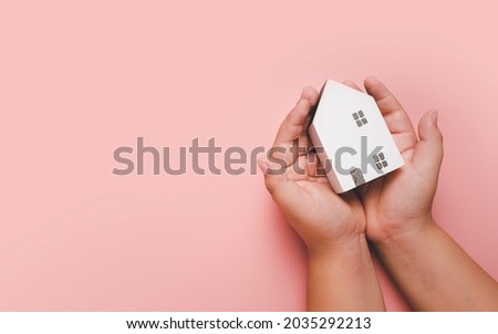 Little boy hand holding white house on pink background, homeless housing and home protecting insurance concept, international day of families, homeschooling, Top view.