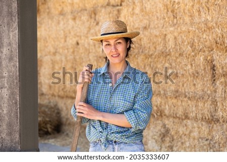 Portrait of positive young asian female farmer standing leaning on pitchfork near straw stack in hayloft, posing during working at farm