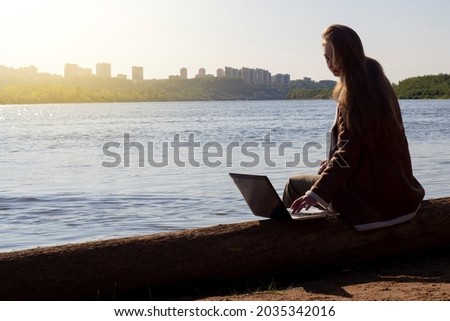 A girl with a laptop is sitting on a log by the river bank in a park outside the city. Concept of freelance, remote work or training. An independent female businessman works in nature in traveling.