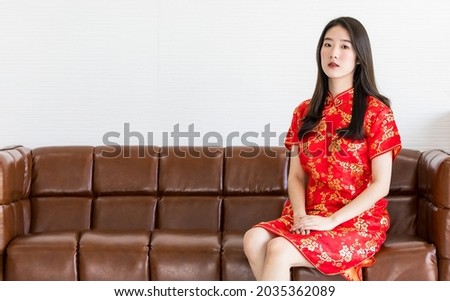A Chinese beautiful woman with long black hairs in red traditional dress is sitting politely on a brown sofa and put hands on her lap Royalty-Free Stock Photo #2035362089