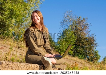 Girl with a laptop is sitting on coastal concrete slabs in a park outside city against background of a forest. Concept of freelance, remote work. Independent female businessman works in nature travel.
