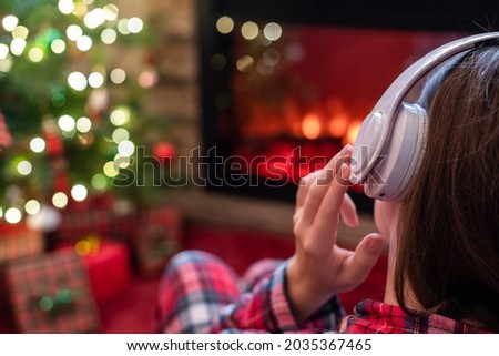 Woman in pajama in headphones sitting and warming at winter evening near fireplace flame and  christmas tree. 