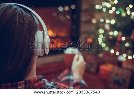 Woman in pajama in headphones with cup of hot cocoa and marshmallow sitting and warming at winter evening near fireplace flame and  christmas tree.
