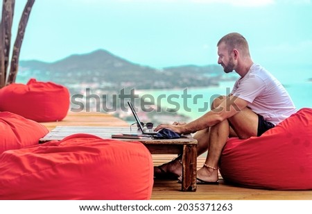 A Caucasian man sitting on a red cushion working remotely with his laptop. There are mountains and the sea in the background Royalty-Free Stock Photo #2035371263