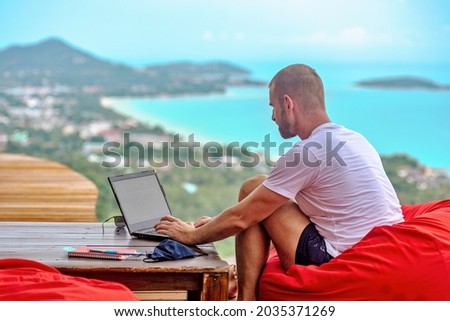 A Caucasian man sitting on a red cushion working remotely with his laptop. There are mountains and the sea in the background Royalty-Free Stock Photo #2035371269
