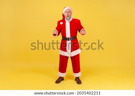 Full length portrait of elderly man with gray beard wearing santa claus costume standing with shocking expression and pointing down with both hands. Indoor studio shot isolated on yellow background.