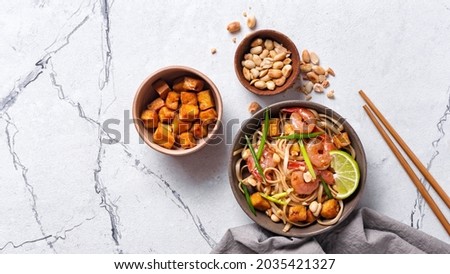 Top view of plate with asian udon noodles, spicy shrimps, fried tofu cheese, peanuts, lime and green onion on white marble background