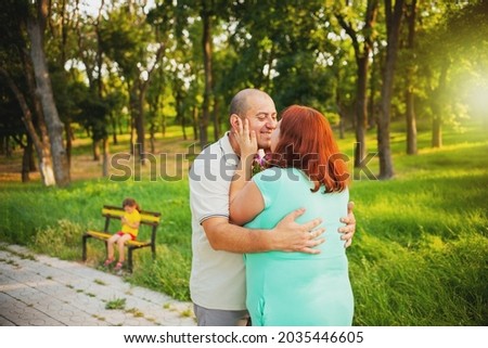 Beautiful loving couple of pregnant woman and her husband standing together on the path in the park embracing and kissing, their little daughter is sitting on a bench.