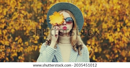 Autumn portrait of beautiful young woman covering her eyes with yellow maple leaves blowing her lips wearing a round hat in a park