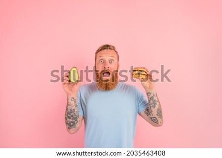 Amazed man with beard and tattoos is undecided if to eat an avocado or an hamburger