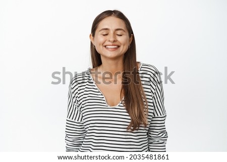 Young dreamy woman stands with closed eyes and happy smile, daydreaming, making wish or thinking of smth, standing against white background.