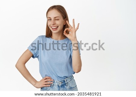 Fine, no problem. Smiling blond woman assure you, show okay OK sign and winking satisfied, praise excellent work, good job, standing in t-shirt over white background.