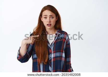 Who me. Ginger girl pointing at her and look with disbelief and surprise, hear something stranger about herself, accused, standing over white background.