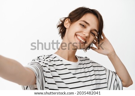 Beautiful brunette woman taking selfie, extend hand, holding camera and posing, standing in t-shirt against white background.