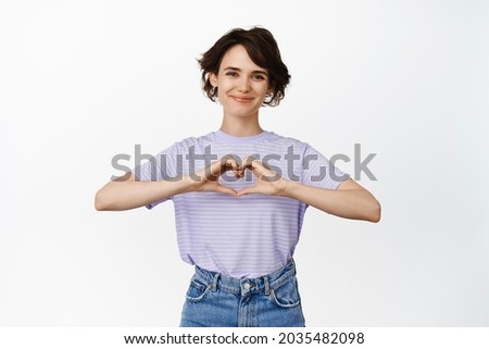 Image of romantic, cute woman showing heart sign and smiling, I love you gesture, sympathy or affection, like smth good, standing over white background.