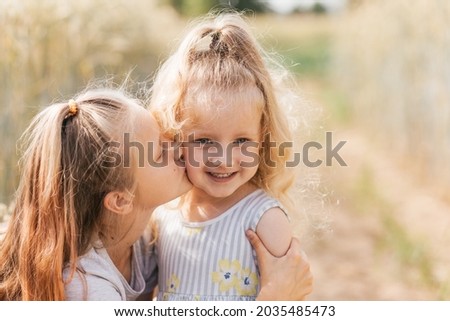 Older sister kisses the cheek of the younger in the summer in nature. Kids hug.