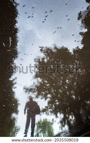Blurred silhouette of reflection of one person walking alone on wet sidewalk of city park on rainy day. Birds are flying across the sky. Abstract photography. Royalty-Free Stock Photo #2035508018