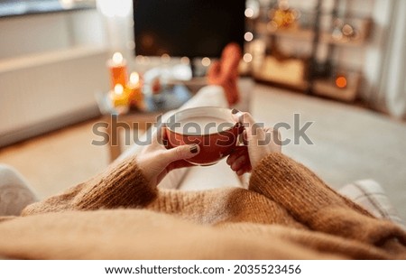 people and leisure concept - hands of young woman watching tv and drinking hot chocolate at cozy home