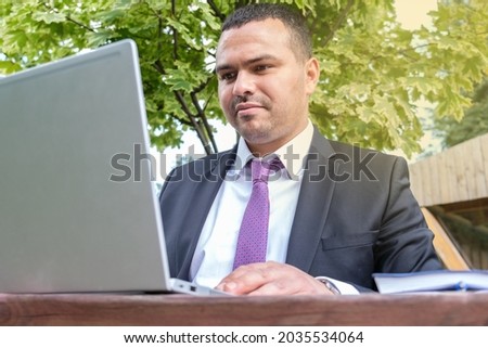 Middle eastern man is typing on a laptop. Serious young man in business suit and tie works on open veranda. Business portrait of leading field manager. Busy man with short hair.